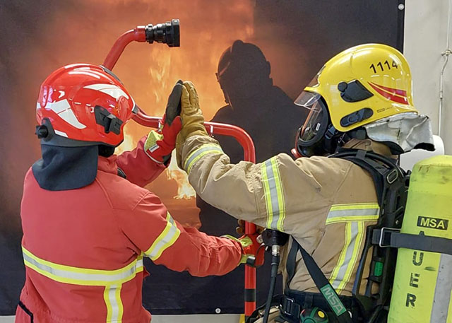 Lähikuva kahdesta palomiehestä takaapäin, kun he nostavat kätensä ylävitoseen; tuote näkyy taustalla.