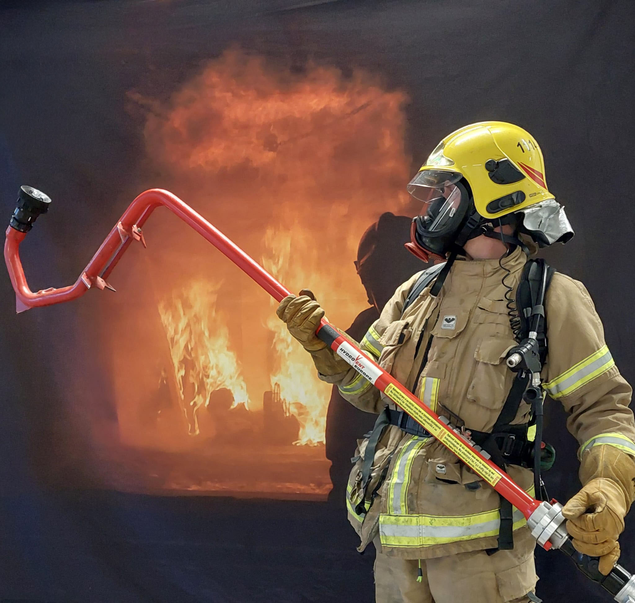 Palomies varusteissaan pitää tuotetta kädessään taustalla palavassa tilassa.